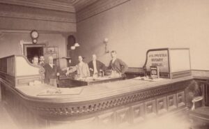Undated photograph of E.W. Churchill (second from left) behind the desk at the Goodman Bank.